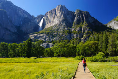 Young woman explores Yosemite Valley - Sentinel / Cook&#39;s Meadow Loop on a wooden boardwalk, with Yosemite Falls in sight, on a sunny day of summer