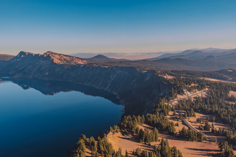 An aerial view of Rim Drive winding around Crater Lake with surrounding mountain peaks and pine trees. Deep dark blue lake reflections in Crater Lake National Park, Oregon, USA.