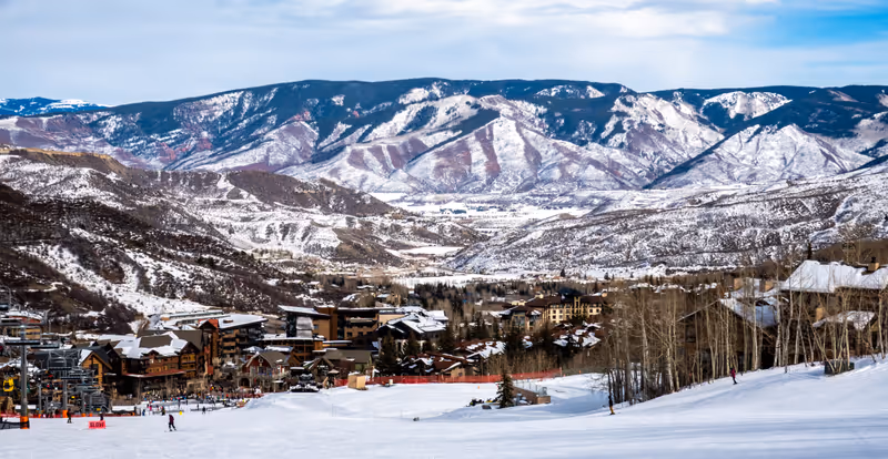 Panoramic view of Snowmass Village, with skiers skiing at the Aspen Snowmass ski resort in the foreground and the Rocky Mountains of Colorado in the background, on a partly cloudy winter day.