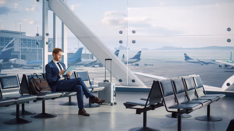 Airport Terminal Flight Wait: Smiling Businessman Uses Digital Tablet Computer for e-Business, Browsing Internet with App. Traveling Entrepreneur Work Online, Sitting in Boarding Lounge of Airline Hub