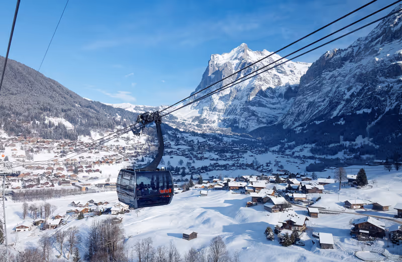 Winter scenery of Grindelwald village on the snowy hillside with Wetterhorn mountain under blue sky in background, viewed from a gondola of Eiger Express cableway, in Berner Oberland, Switzerland