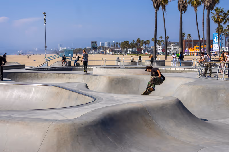 Los Angeles, California USA - 3 20 2024: a gorgeous spring landscape at Venice Beach Skatepark at Venice Beach with young men riding skateboards and people watching, palm trees, buildings and blue sky