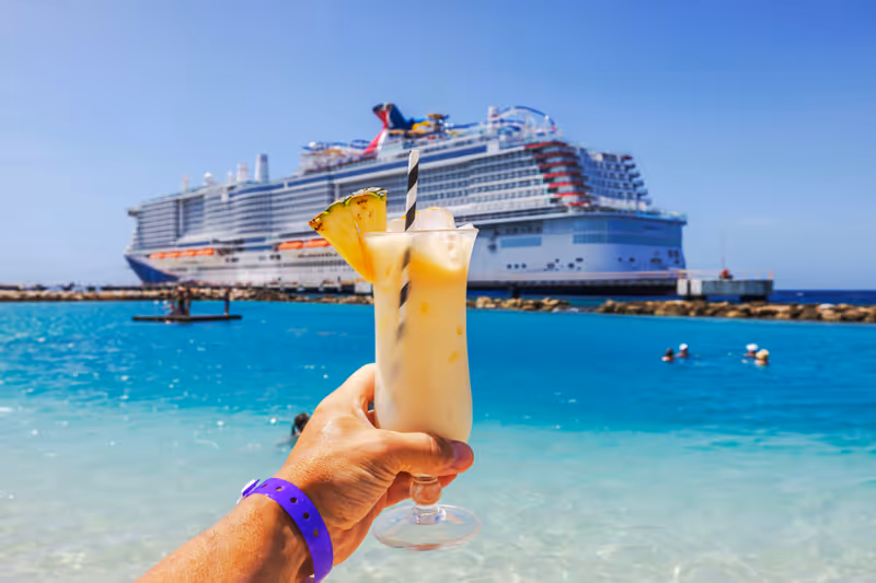 Close-up view of hand holding glass of alcoholic cocktail on Caribbean beach with cruise ship in background.