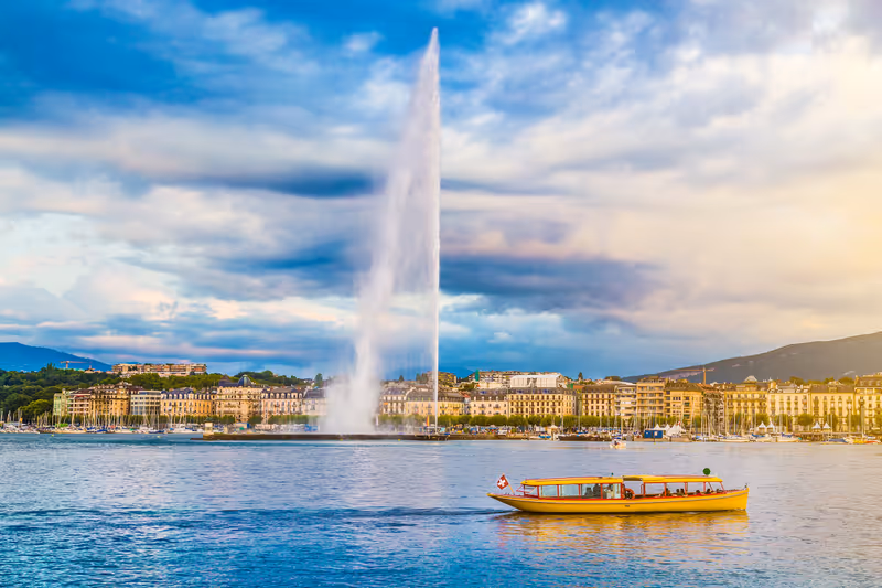 Panoramic view of Geneva skyline with famous Jet d'Eau fountain and traditional boat at harbor district in beautiful evening light at sunset, Canton of Geneva, Switzerland